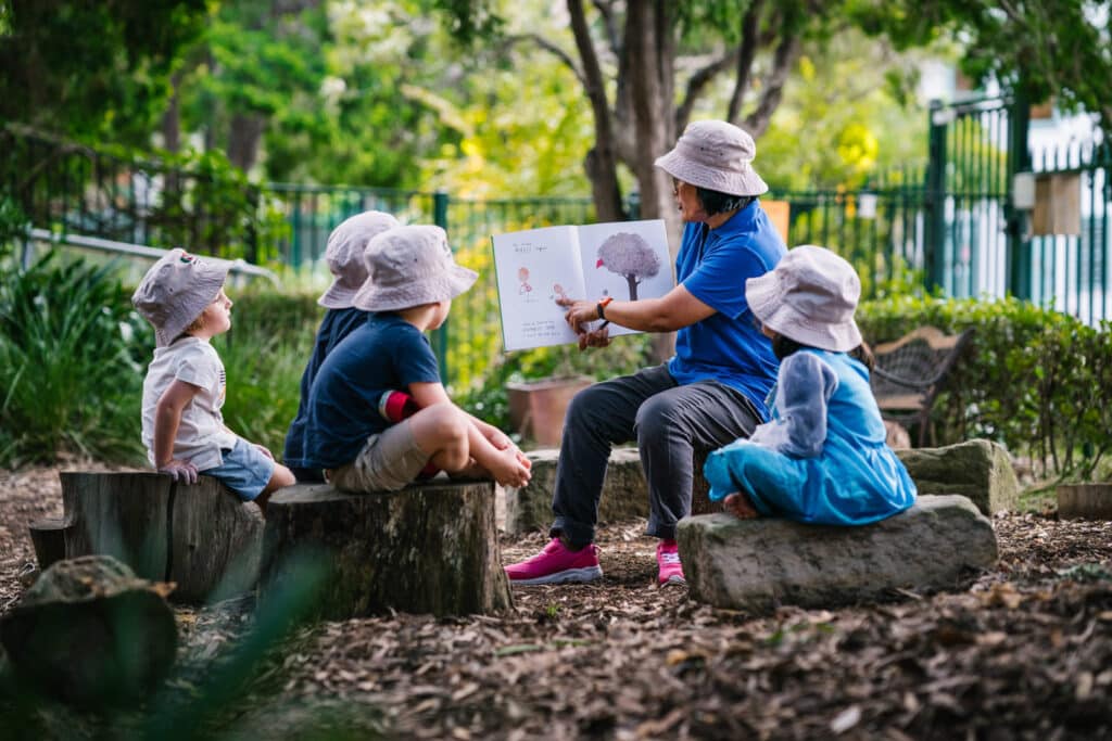 KU Rushcutters Bay childcare educator reading a story to children outdoors