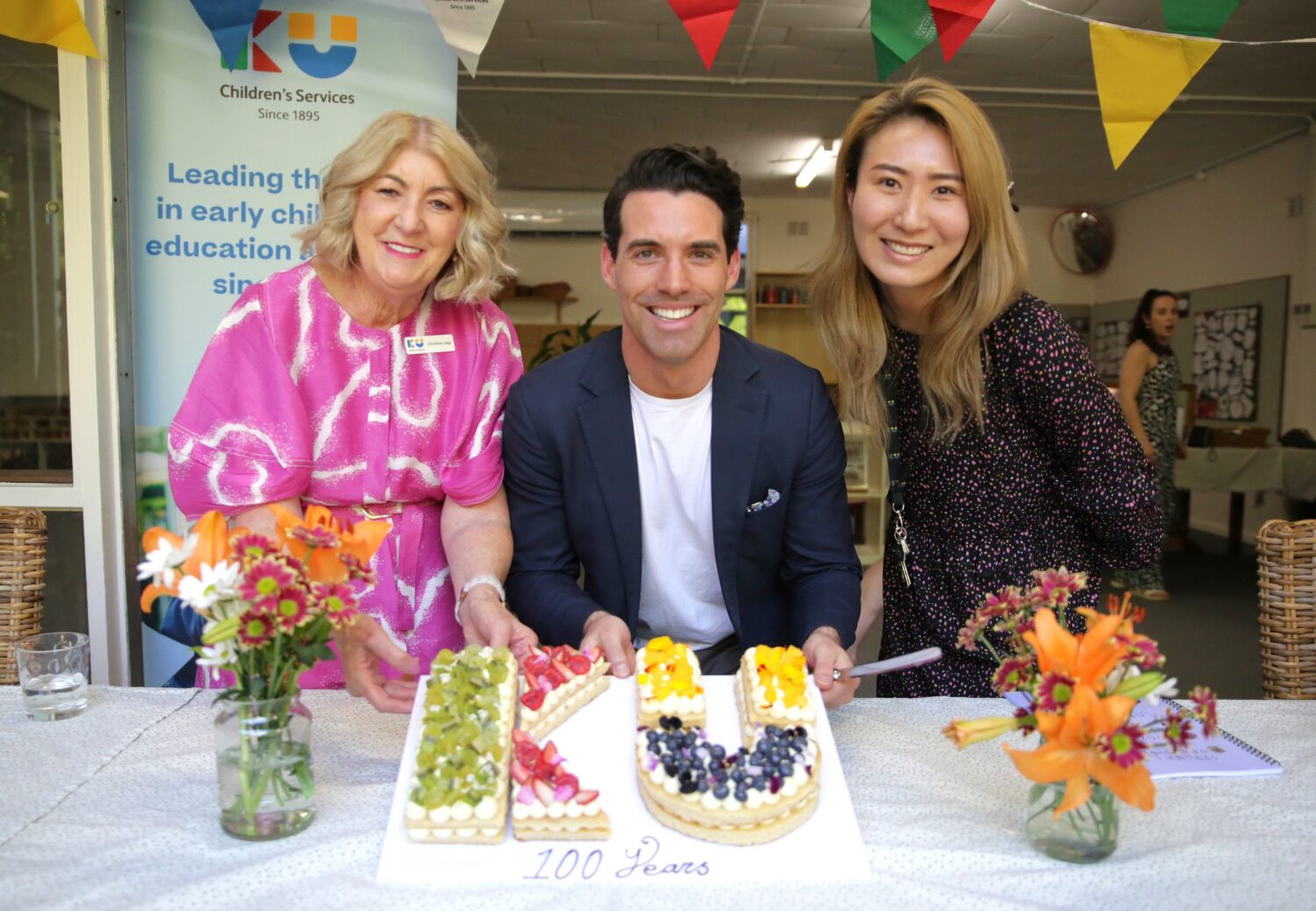 KU Sunbeam Preschool celebrates 100th anniversary 6 Chris Legg, Zann Maxwell, Sarah Kim, posing for a photograph with KU shaped cake.